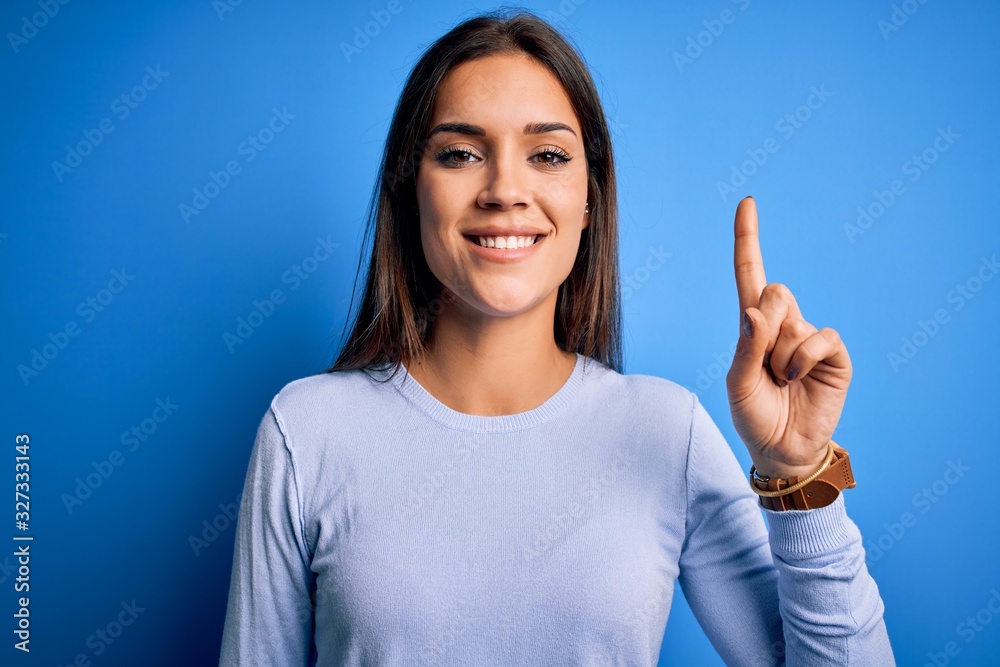 Young beautiful brunette woman wearing casual sweater standing over blue background showing and pointing up with finger number one while smiling confident and happy.