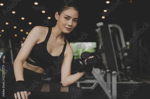 athletic asian slim body woman smiling looking at camera and exercise with dumbbell in fitness gym in background, bodybuilder, healthy lifestyle, exercise fitness, workout and sport training concept