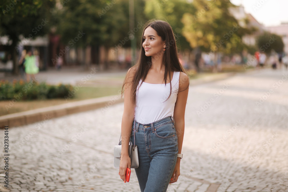 Attractive young woman walking in centre of city in summer time. Beautiful brunette in white shirt. Smile