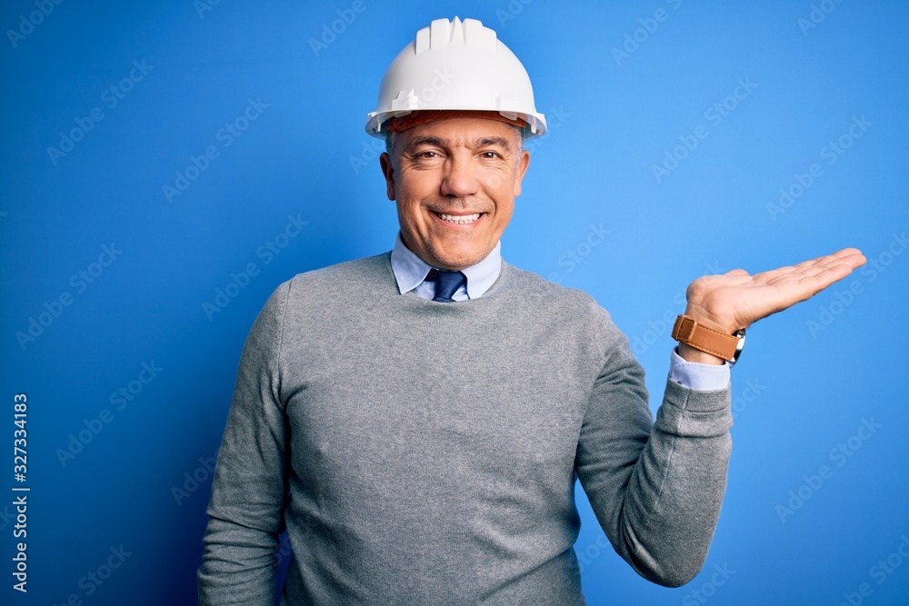 Middle age handsome grey-haired engineer man wearing safety helmet over blue background smiling cheerful presenting and pointing with palm of hand looking at the camera.