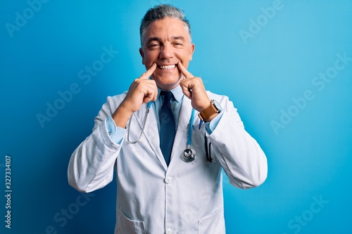Middle age handsome grey-haired doctor man wearing coat and blue stethoscope Smiling with open mouth, fingers pointing and forcing cheerful smile
