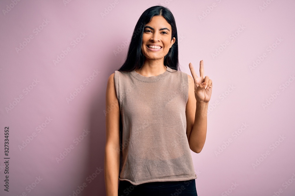 Young beautiful hispanic fashion woman wearing sweater over pink background smiling with happy face winking at the camera doing victory sign. Number two. Young beautiful hispanic fashion woman wearing sweater over pink background smiling with happy face winking at the camera doing victory sign. Number two.