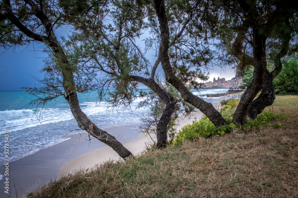 Seafront of Tyrrhenian Sea. View of the Sicilian city of Cefalu through trees growing on the shore