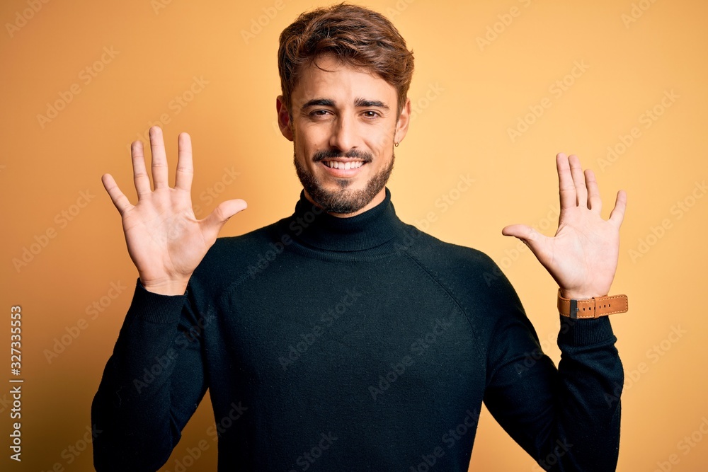 Young handsome man with beard wearing turtleneck sweater standing over yellow background showing and pointing up with fingers number ten while smiling confident and happy. Young handsome man with beard wearing turtleneck sweater standing over yellow background showing and pointing up with fingers number ten while smiling confident and happy.