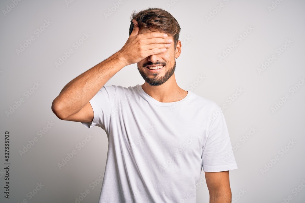 Young handsome man with beard wearing casual t-shirt standing over white background smiling and laughing with hand on face covering eyes for surprise. Blind concept. Young handsome man with beard wearing casual t-shirt standing over white background smiling and laughing with hand on face covering eyes for surprise. Blind concept.