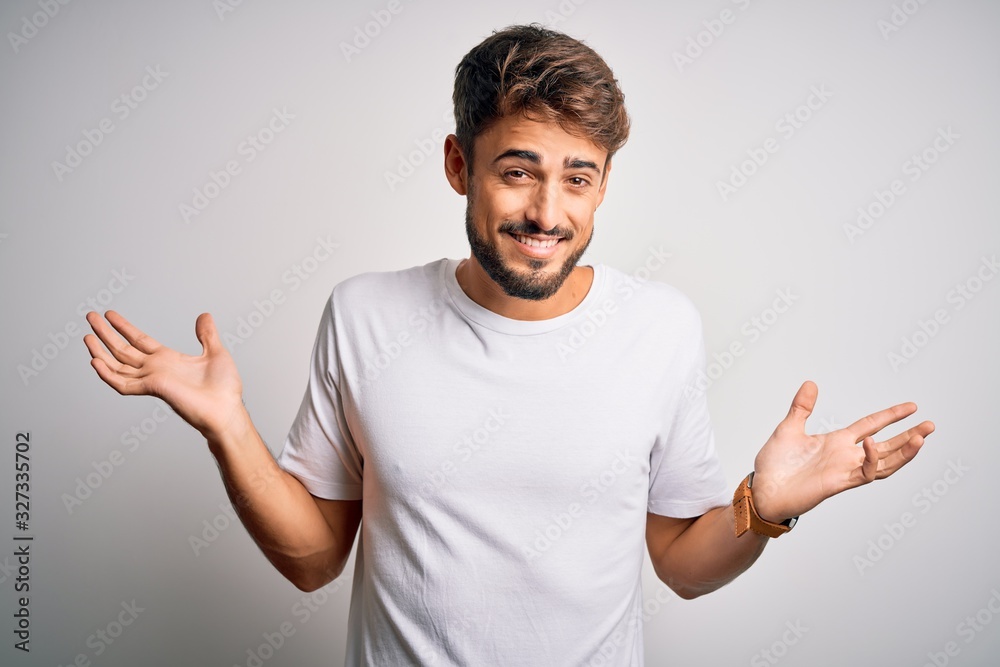 Young handsome man with beard wearing casual t-shirt standing over white background clueless and confused expression with arms and hands raised. Doubt concept. Young handsome man with beard wearing casual t-shirt standing over white background clueless and confused expression with arms and hands raised. Doubt concept.