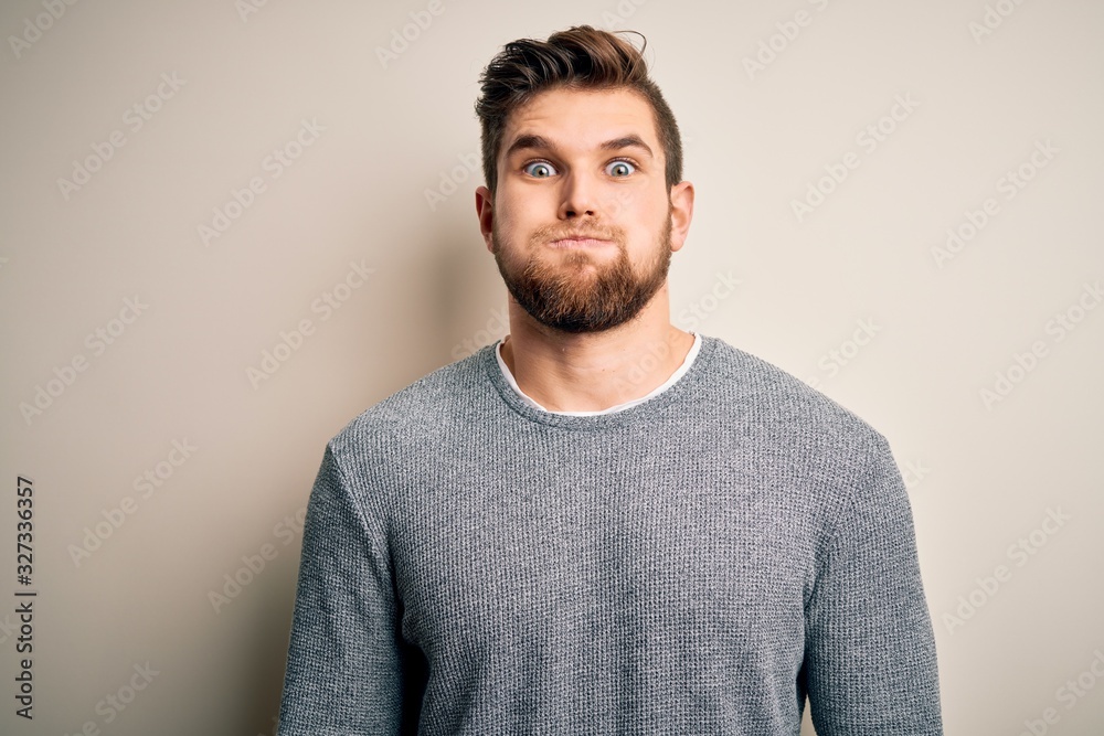 Young handsome blond man with beard and blue eyes wearing casual sweater puffing cheeks with funny face. Mouth inflated with air, crazy expression. Young handsome blond man with beard and blue eyes wearing casual sweater puffing cheeks with funny face. Mouth inflated with air, crazy expression.