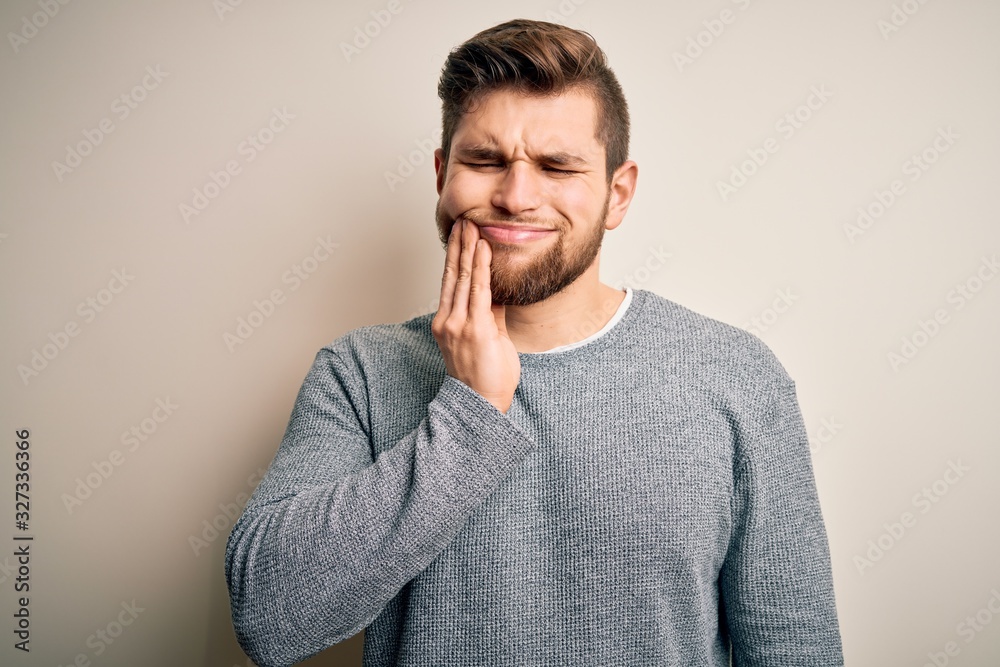 Young handsome blond man with beard and blue eyes wearing casual sweater touching mouth with hand with painful expression because of toothache or dental illness on teeth. Dentist Young handsome blond man with beard and blue eyes wearing casual sweater touching mouth with hand with painful expression because of toothache or dental illness on teeth. Dentist