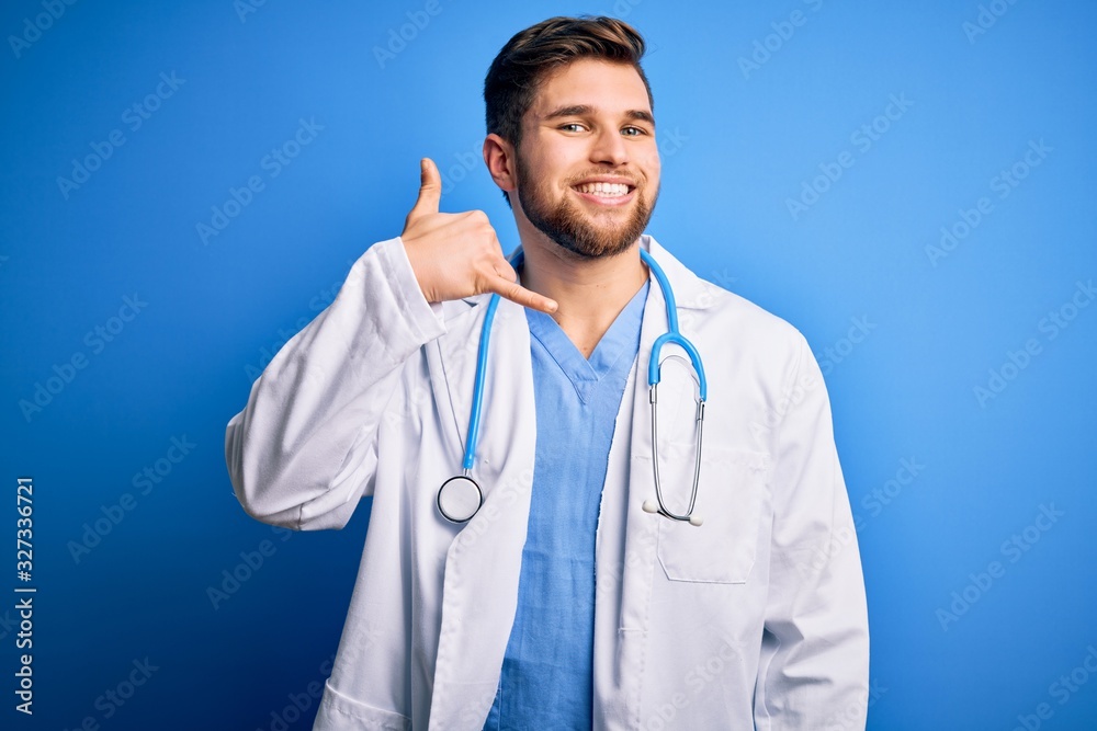 Young blond doctor man with beard and blue eyes wearing white coat and stethoscope smiling doing phone gesture with hand and fingers like talking on the telephone. Communicating concepts. Young blond doctor man with beard and blue eyes wearing white coat and stethoscope smiling doing phone gesture with hand and fingers like talking on the telephone. Communicating concepts.