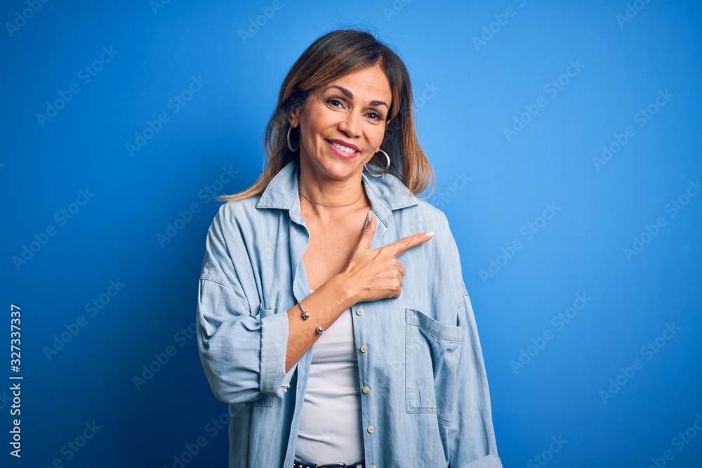 Middle age beautiful woman wearing casual shirt standing over isolated blue background cheerful with a smile on face pointing with hand and finger up to the side with happy and natural expression Middle age beautiful woman wearing casual shirt standing over isolated blue background cheerful with a smile on face pointing with hand and finger up to the side with happy and natural expression