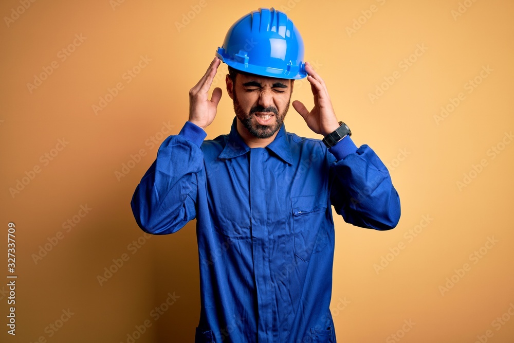Mechanic man with beard wearing blue uniform and safety helmet over yellow background with hand on headache because stress. Suffering migraine.