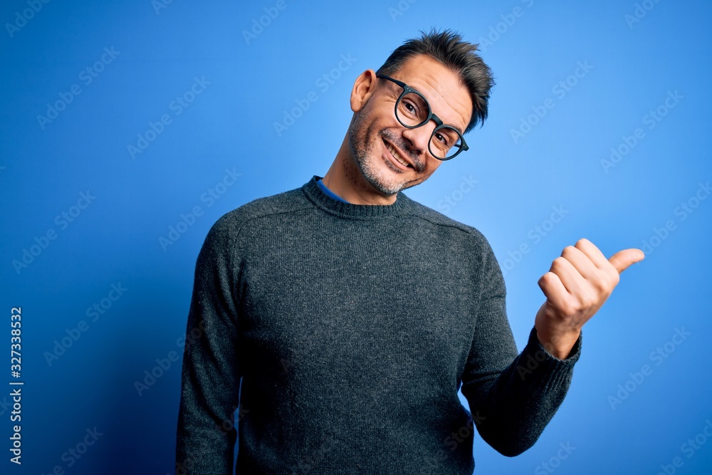 Young handsome man wearing casual sweater and glasses standing over blue background smiling with happy face looking and pointing to the side with thumb up. Young handsome man wearing casual sweater and glasses standing over blue background smiling with happy face looking and pointing to the side with thumb up.