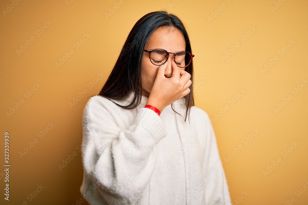 Young beautiful asian woman wearing casual sweater and glasses over yellow background tired rubbing nose and eyes feeling fatigue and headache. Stress and frustration concept.
