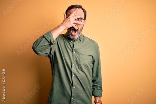 Middle age hoary man wearing casual green shirt and glasses over isolated yellow background peeking in shock covering face and eyes with hand  looking through fingers with embarrassed expression.