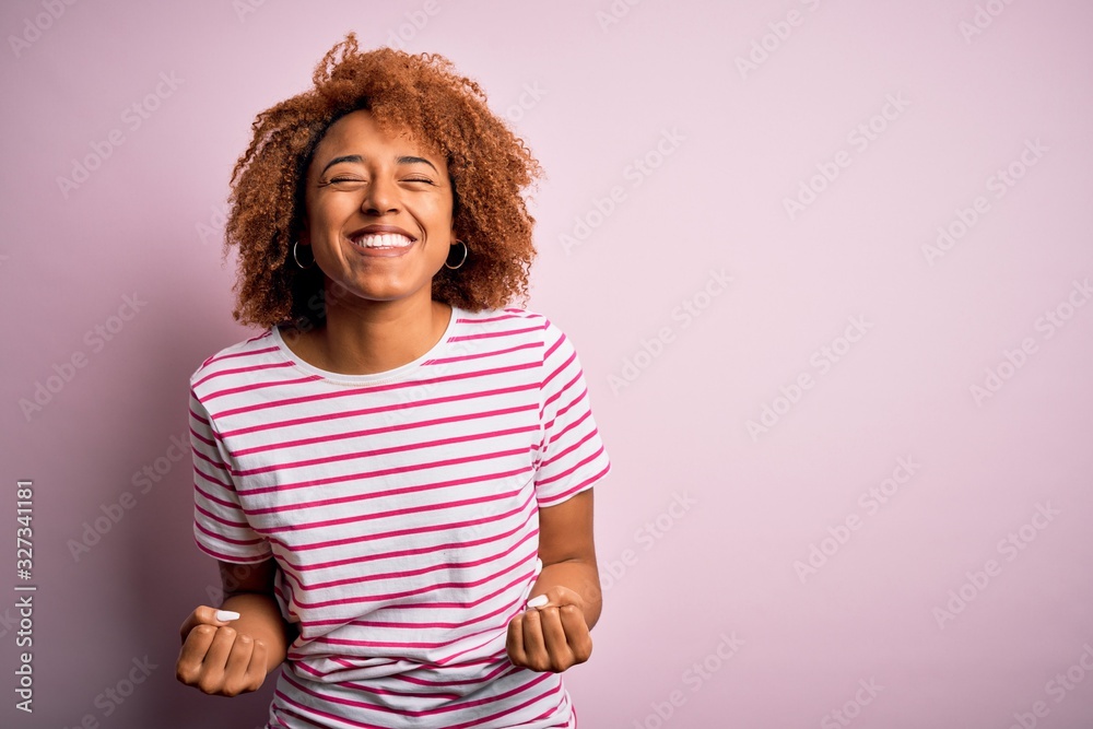 Young beautiful African American afro woman with curly hair wearing casual striped t-shirt very happy and excited doing winner gesture with arms raised, smiling and screaming for success. Celebratio
