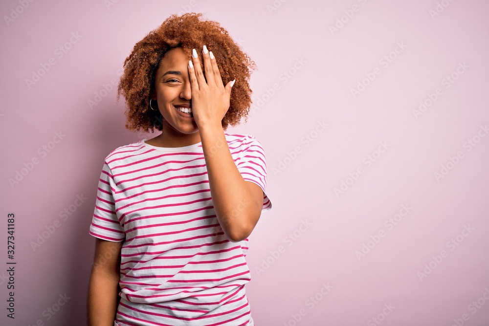 Young beautiful African American afro woman with curly hair wearing casual striped t-shirt covering one eye with hand, confident smile on face and surprise emotion.