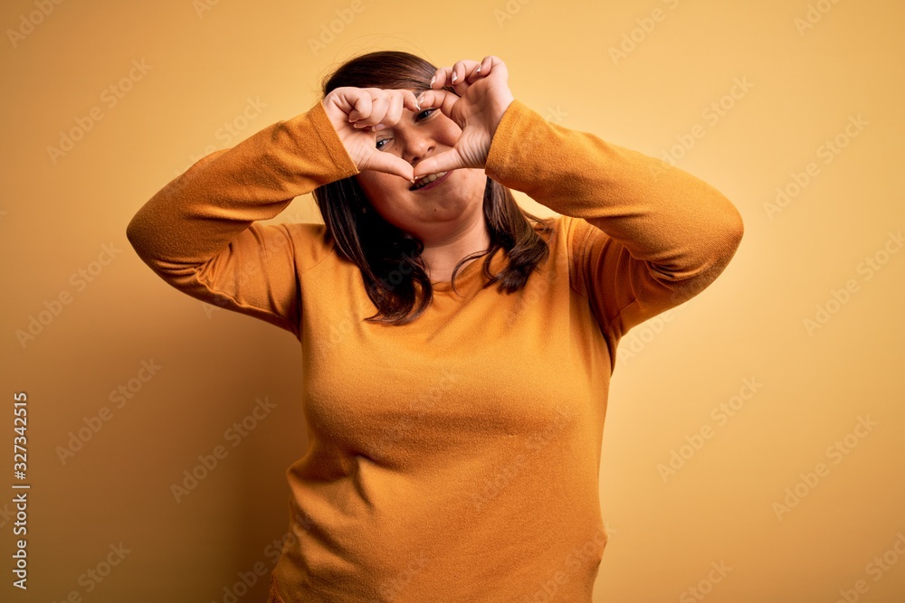 Beautiful brunette plus size woman wearing casual sweater over isolated yellow background Doing heart shape with hand and fingers smiling looking through sign Beautiful brunette plus size woman wearing casual sweater over isolated yellow background Doing heart shape with hand and fingers smiling looking through sign