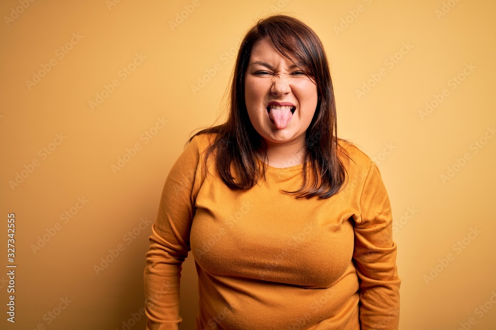 Obraz premium Beautiful brunette plus size woman wearing casual sweater over isolated yellow background sticking tongue out happy with funny expression. Emotion concept.
