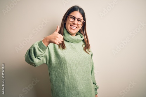 Young beautiful woman wearing casual sweater standing over isolated white background doing happy thumbs up gesture with hand. Approving expression looking at the camera showing success. © Krakenimages.com
