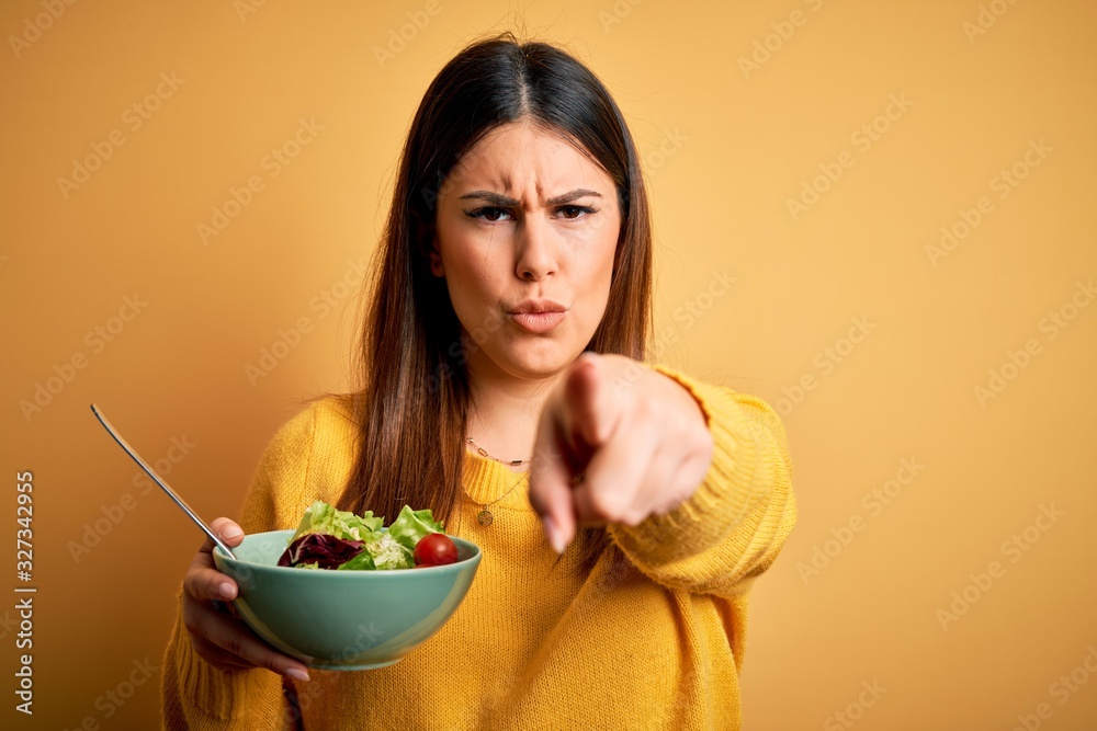 Young beautiful woman eating healthy fresh salad over yellow background pointing with finger to the camera and to you, hand sign, positive and confident gesture from the front Young beautiful woman eating healthy fresh salad over yellow background pointing with finger to the camera and to you, hand sign, positive and confident gesture from the front