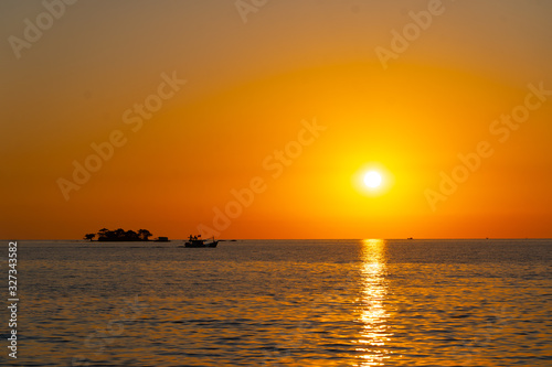 orange tropical sunset with little island and fisherman boat on horizon line