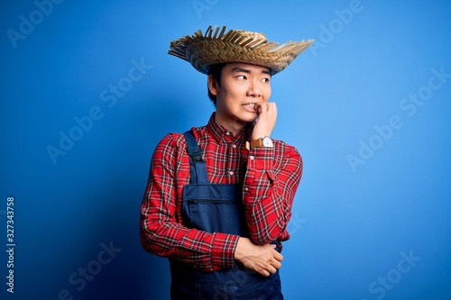 Young handsome chinese farmer man wearing apron and straw hat over blue background looking stressed and nervous with hands on mouth biting nails. Anxiety problem.