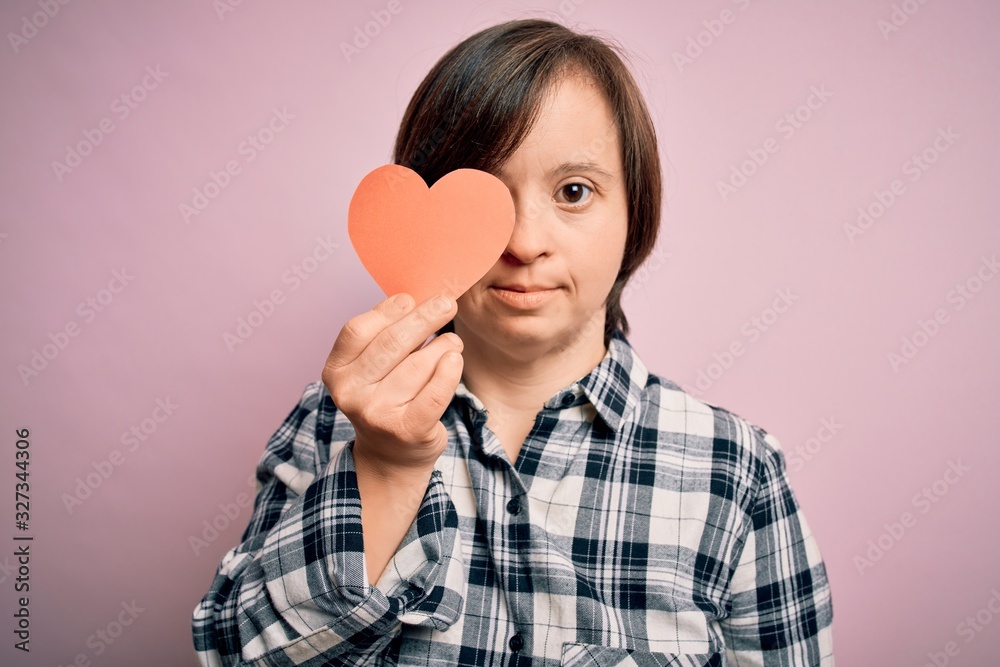 Young down syndrome romantic woman holding red heart paper shape over pink background with a confident expression on smart face thinking serious Young down syndrome romantic woman holding red heart paper shape over pink background with a confident expression on smart face thinking serious