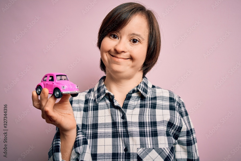 Young down syndrome woman holding small car as driving license insurance over pink background with a happy face standing and smiling with a confident smile showing teeth