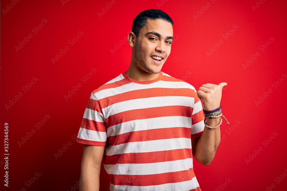 Young brazilian man wearing casual striped t-shirt standing over isolated red background smiling with happy face looking and pointing to the side with thumb up. Young brazilian man wearing casual striped t-shirt standing over isolated red background smiling with happy face looking and pointing to the side with thumb up.
