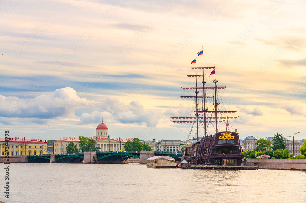 Obraz premium Cityscape of Saint Petersburg Leningrad with bascule Exchange Bridge, sailboat wooden ship with mast moored anchored on water of Neva river, evening dramatic orange sky background, Russia