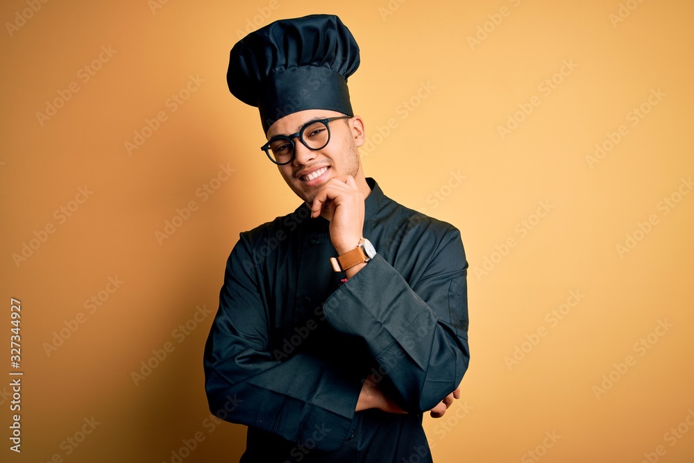 Young brazilian chef man wearing cooker uniform and hat over isolated yellow background looking confident at the camera smiling with crossed arms and hand raised on chin. Thinking positive. Young brazilian chef man wearing cooker uniform and hat over isolated yellow background looking confident at the camera smiling with crossed arms and hand raised on chin. Thinking positive.