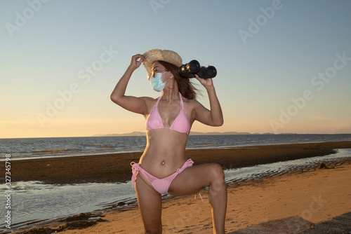 An senior Asian woman dressed in a pink swimsuit and has a nose mask. Protect Virus COVID-19 and PM2.5 protection. Use binoculars to looking.