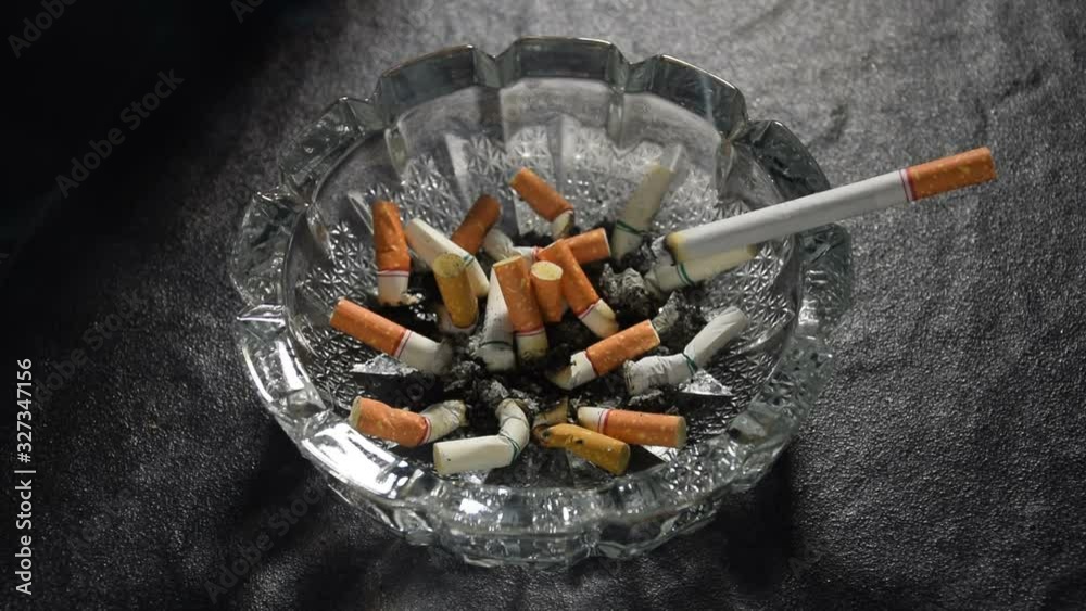 Lit cigarette with smoke lying on an ashtray filled with cigarette butts and ashes . Lit cigarette with smoke lying on an ashtray filled with cigarette butts and ashes .