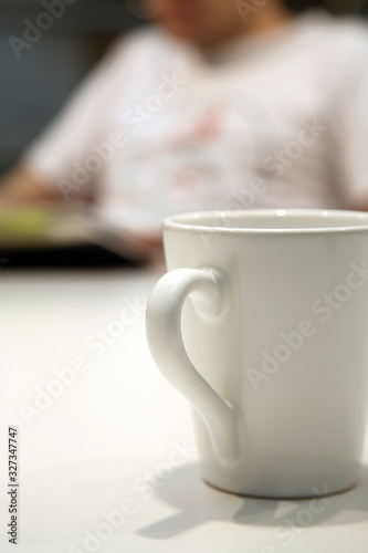 Close up shot of a white coffee mug, with an out of focus person in the background, reading a book