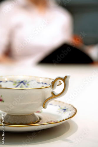 Close up shot of an old fashioned china tea cup and saucer, with an out of focus person in the background, reading a book