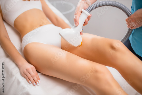 young slender, slim woman get epilation procedure in beauty salon. attractive lady sit in underwear while professional woman applying special ointment and use special machine for hair removal