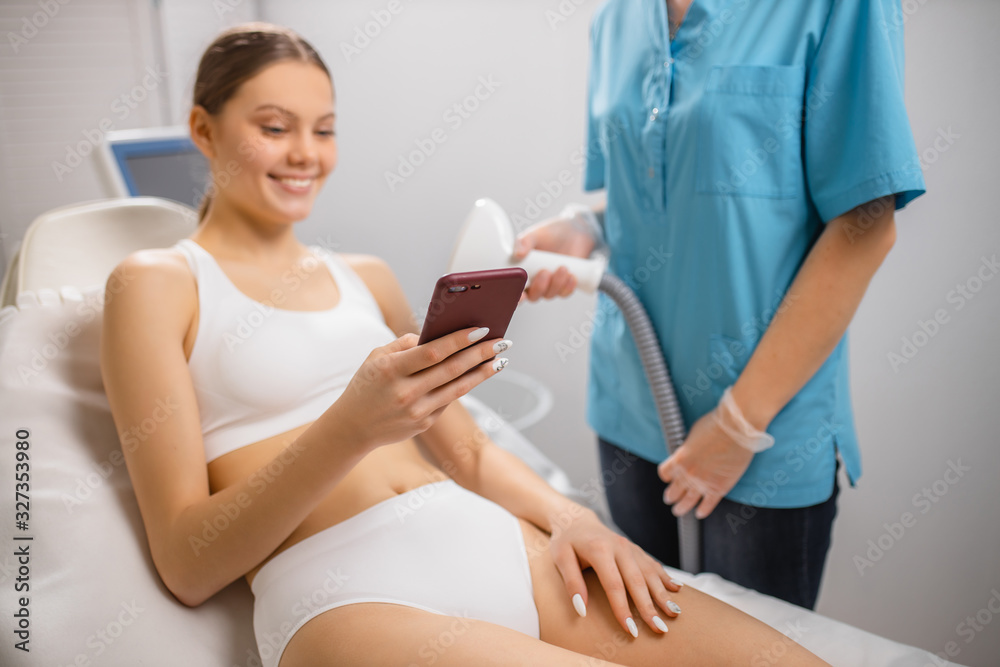young caucasian woman at laser epilation procedure, use smartphone while getting hair removal procedure in beauty salon. hairless, hair removal, epilation, depilation concept young caucasian woman at laser epilation procedure, use smartphone while getting hair removal procedure in beauty salon. hairless, hair removal, epilation, depilation concept