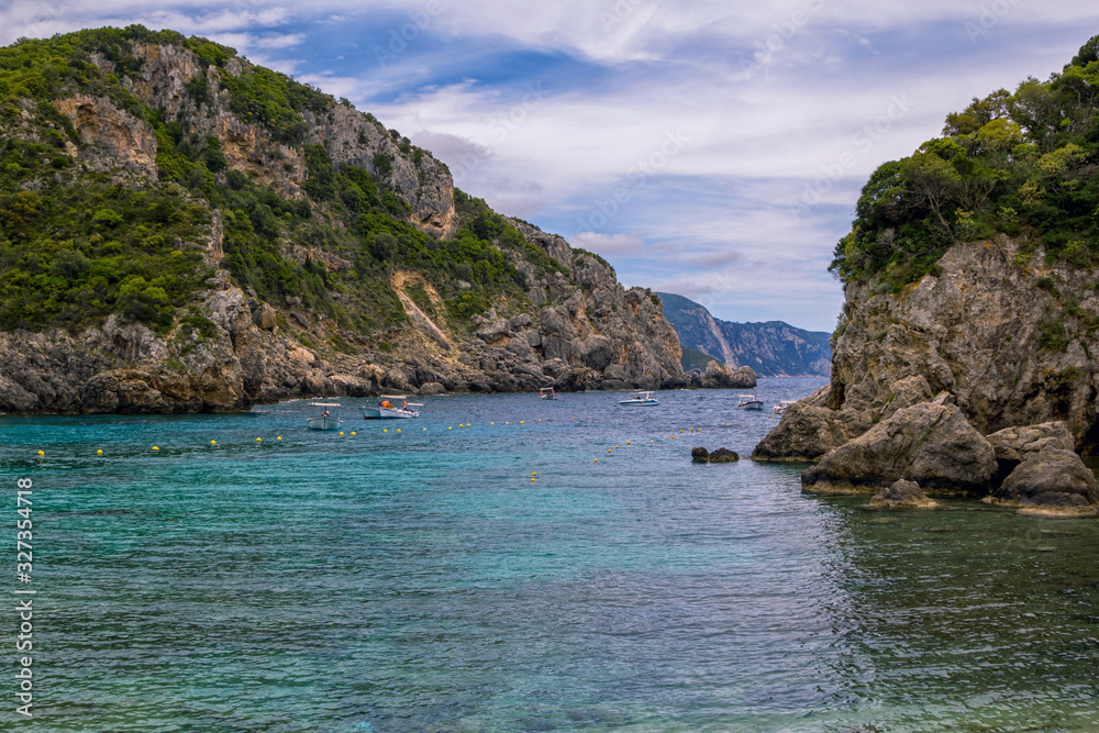 Seascape – lagoon with turquoise water, mountain with cliffs, green trees, bushes, rocks in a blue water and fishers boats. Corfu Island, Greece. 