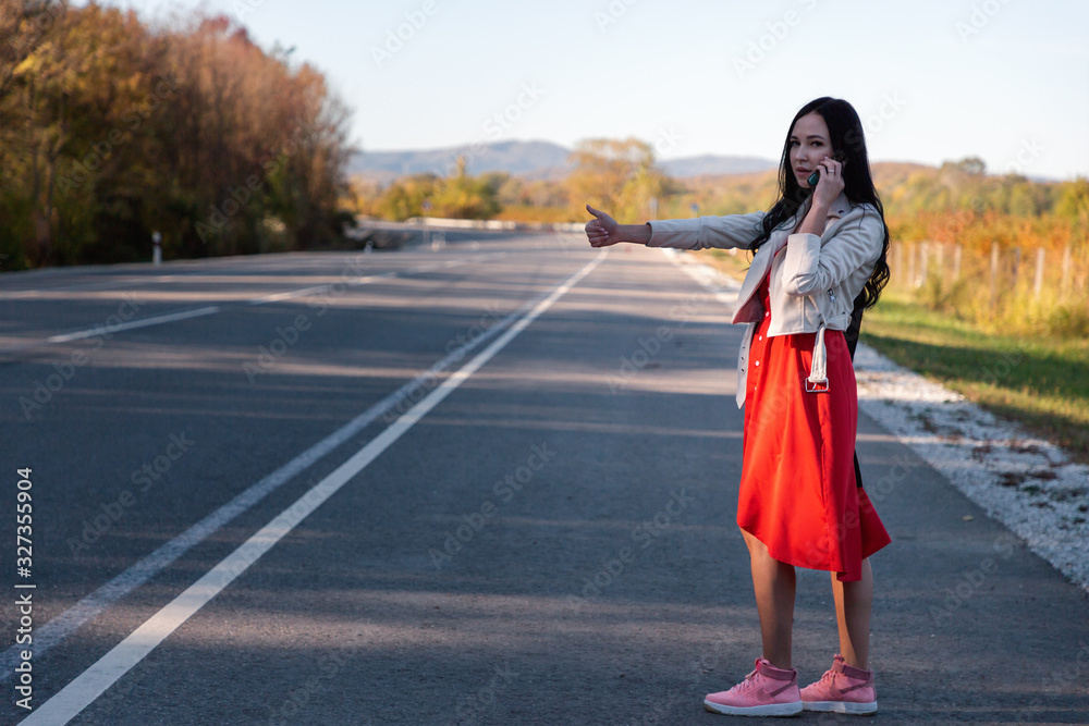 A young beautiful brunette woman in a red dress hitchhiking and calling on the phone. In the background, the road goes into the distance and the trees. The concept of hitchhiking