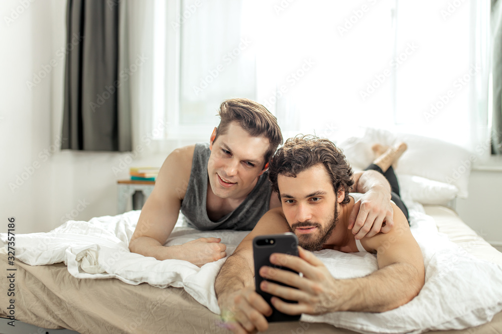 cute homosexual caucasian couple taking selfie on bed at home. two young men in domestic clothes, look at screen of mobile phone and smile, gays in love cute homosexual caucasian couple taking selfie on bed at home. two young men in domestic clothes, look at screen of mobile phone and smile, gays in love