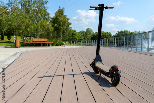 An electric black scooter stands on the bandwagon on the street. City park with wooden flooring along the promenade with railings. Sunny summer day. Modern city transport. photo