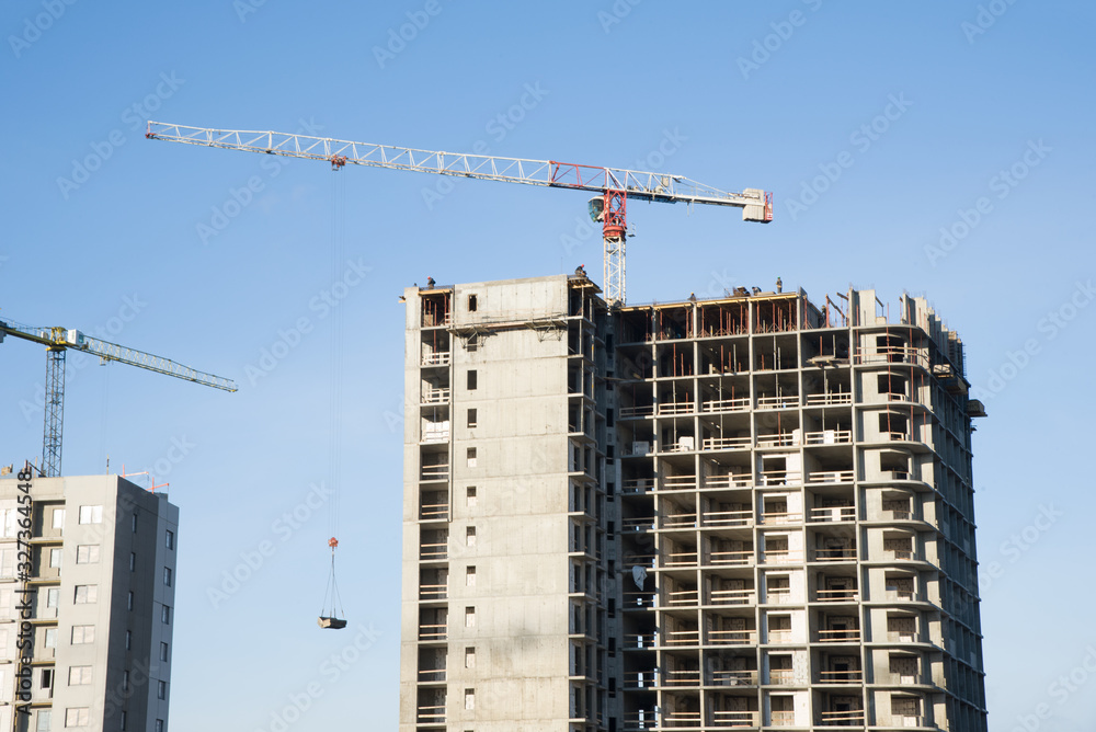 Tower cranes working at construction site against blue sky. Crane lifting a concrete bucket. Construction process of the new residential buildings. Transportation blocks and pouring of the cement mix Tower cranes working at construction site against blue sky. Crane lifting a concrete bucket. Construction process of the new residential buildings. Transportation blocks and pouring of the cement mix