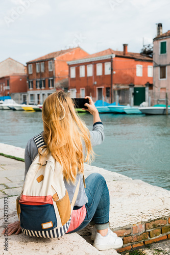 Young female traveler by the canal in Murano in Venice, Italy taking a photo on her smartphone