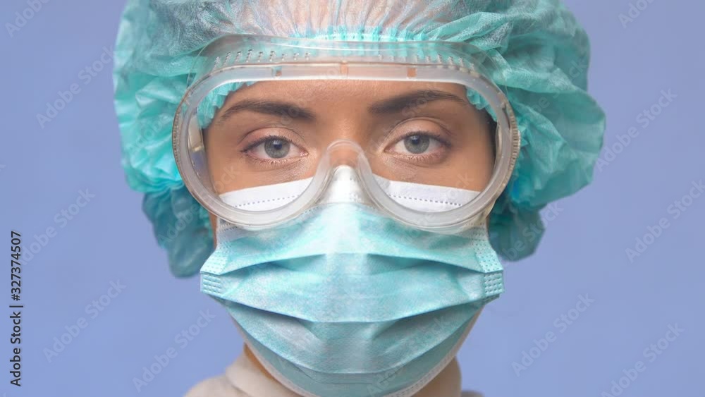 woman doctor in protective medical uniform, goggles and a face mask on a blue medical background looks at the camera with a serious look, close-up portrait. Novel Coronavirus (2019-nCoV), COVID-19