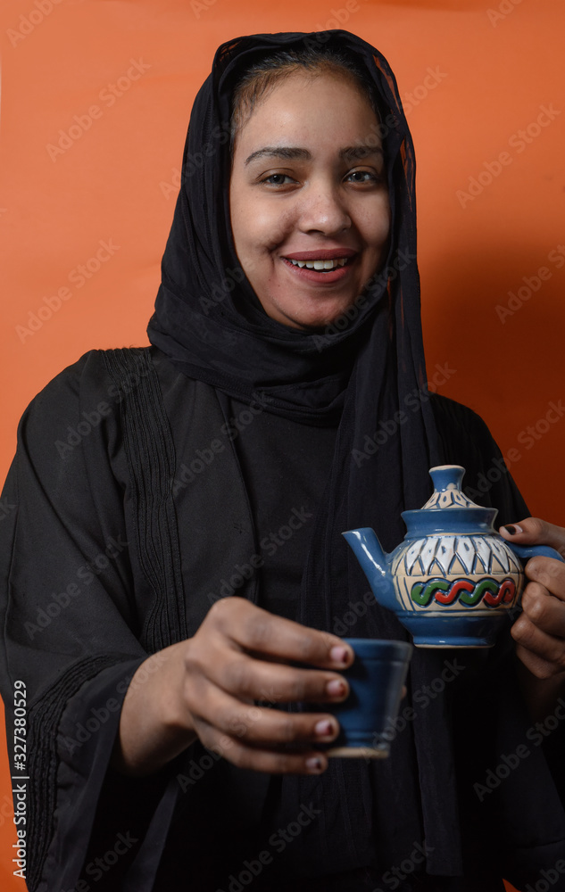 Arabic girl wearing Hijab pouring Tea into tea cup over on orange  background , girl in Arabic Traditional Abbaya