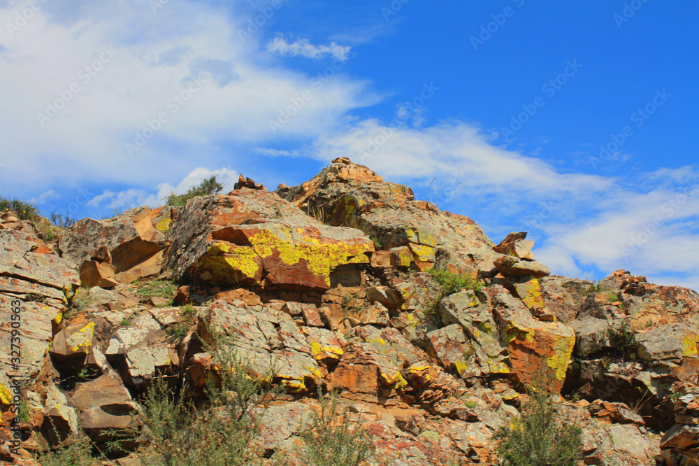 Natural stone wall, a mighty rock with moss, a huge mountain on a background of blue sky with clouds. Red and sand colored cliffs view from below. Wildlife and hiking. Photo from hiking