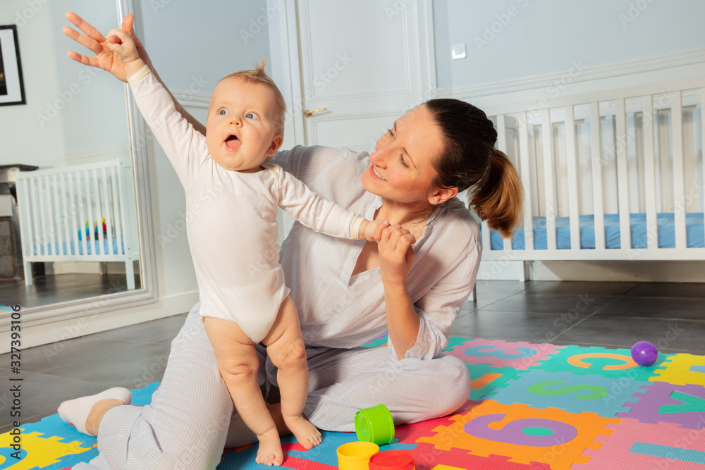 Happy mother play with toddler baby boy in the nursery helping him to stand and lift hands Happy mother play with toddler baby boy in the nursery helping him to stand and lift hands