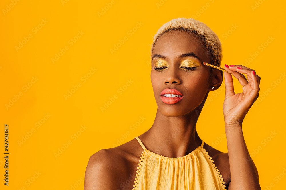 Portrait of a beautiful young woman applying yellow eyeshadow make up to her eyes, looking down - obrazy, fototapety, plakaty 