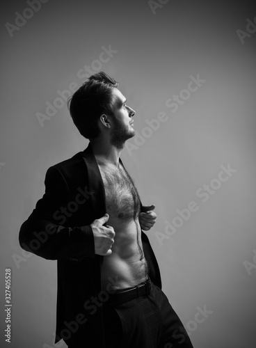 Male with cool hairstyle, in black suit on naked body. He opened his jacket and posing sideways on gray background. Close up
