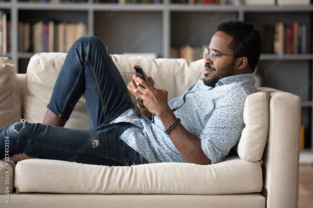 Young african american man lying on cozy sofa, playing games. - obrazy, fototapety, plakaty 